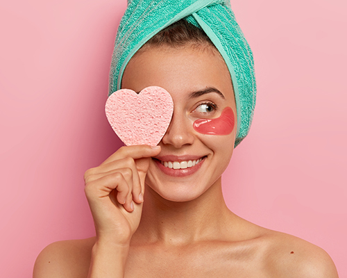 Mujer con una toalla verde en la cabeza y antiojeras se tapa un ojo con una esponja en forma de corazón mientras sonríe